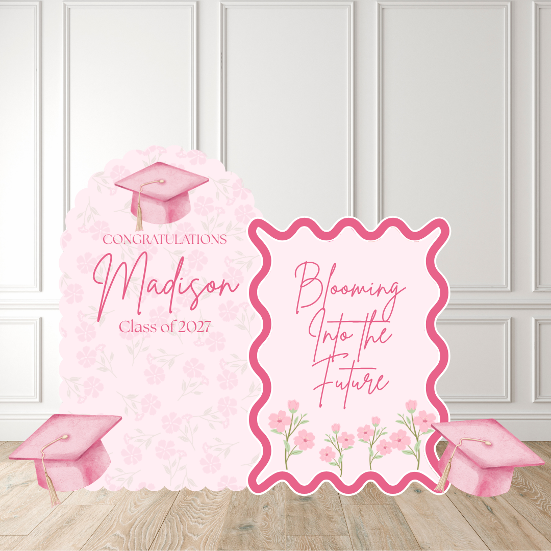 Pink graduation party backdrop set featuring a large arch sign that reads 'Congratulations Madison Class of 2027' with a graduation cap illustration, a smaller decorative sign reading 'Blooming Into the Future' with floral accents, and two small graduation cap props on a light wood floor against a white paneled wall.
