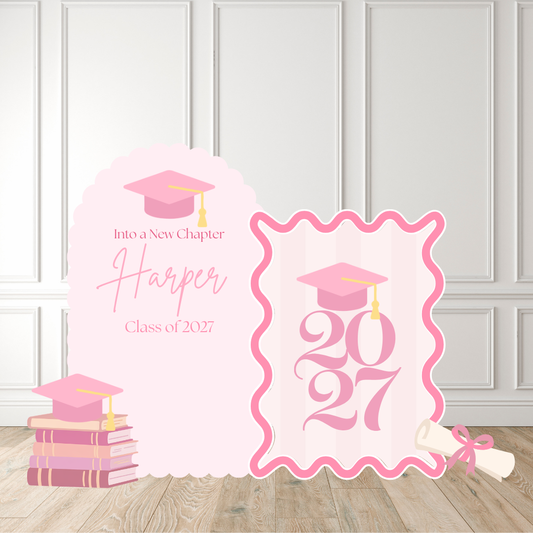 Pink graduation-themed party backdrop standee set on a light wood floor against a white paneled wall. The set includes a tall arch-shaped panel reading 'Into a New Chapter Harper Class of 2027' with a pink graduation cap and books, and a scalloped rectangular panel with '2027' and a graduation cap, plus a rolled diploma with a pink ribbon.