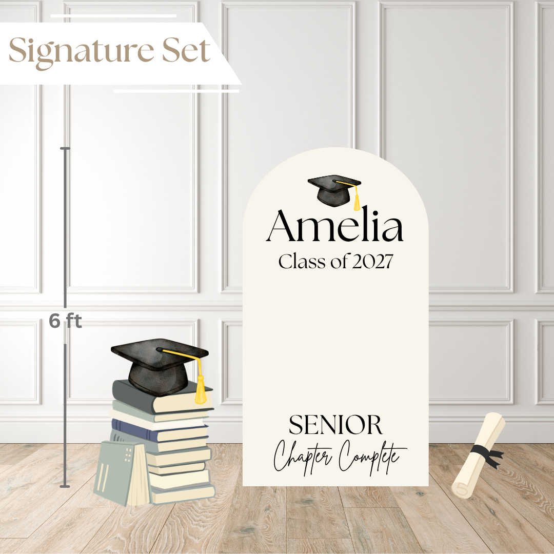 Graduation-themed decorative sign set labeled 'Signature Set' on a light wood floor against a white paneled wall. The set includes a large arched sign reading 'Amelia Class of 2027' and 'Senior Chapter Complete,' plus smaller props: a stack of books with a graduation cap on top, a 6 ft height marker, and a rolled diploma.
