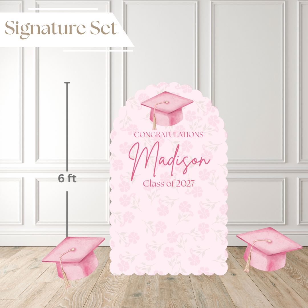 A pink graduation party backdrop set shown in a room scene with wood flooring and white paneled walls. The main product is a tall scalloped arch sign reading “Congratulations Madison Class of 2027” with a pink graduation cap illustration, plus two matching cap props on the floor.