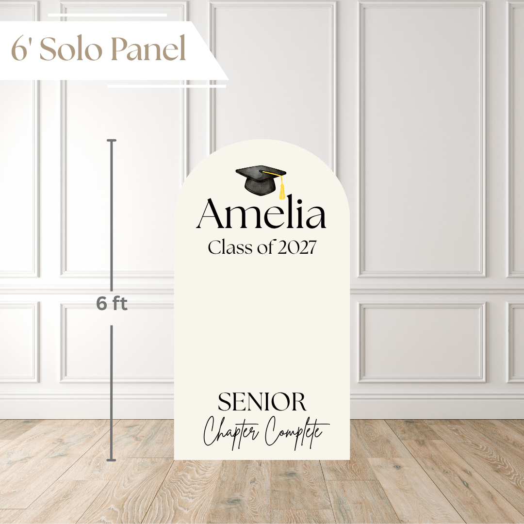 A 6-foot tall graduation solo panel displayed in a room setting with white paneled walls and light wood flooring. The panel is cream-colored with a black arched top, featuring customizable text 'Amelia Class of 2027' and 'SENIOR Chapter Complete' along with a small graduation cap illustration.