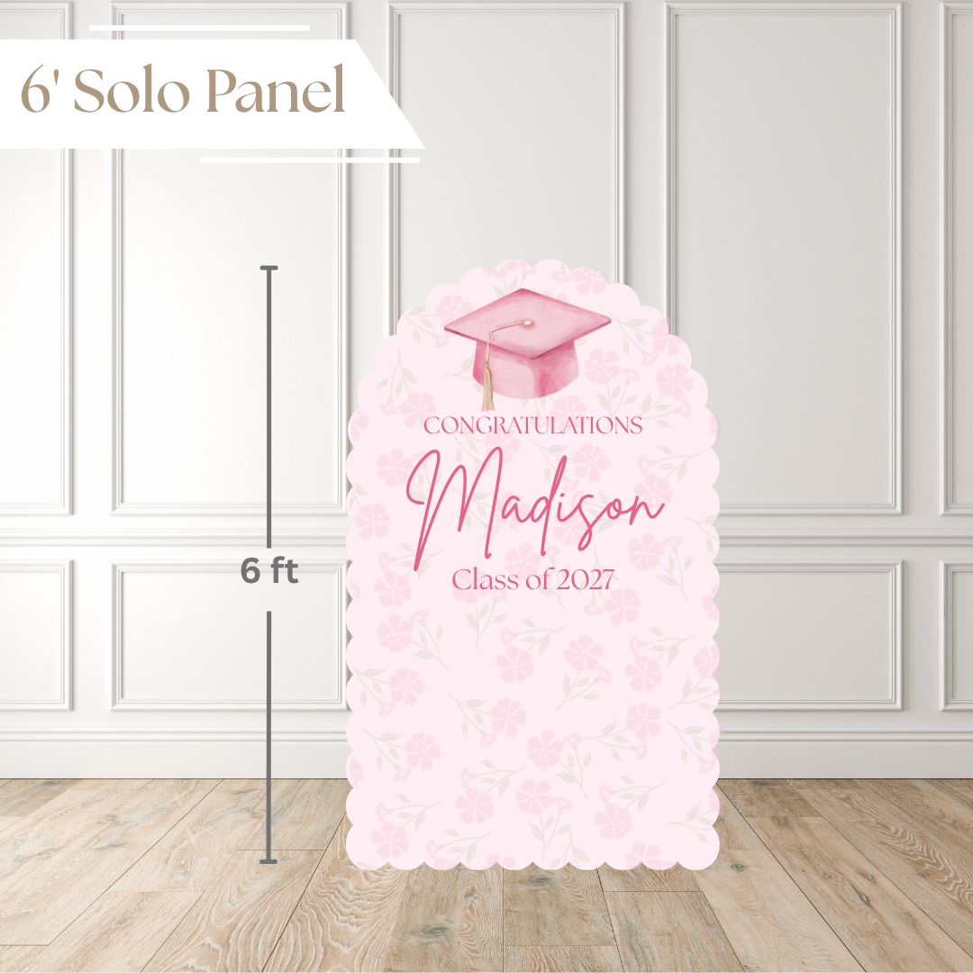 A 6-foot tall graduation solo panel with a pink floral pattern and a watercolor graduation cap illustration. The panel features customizable text reading 'CONGRATULATIONS Madison Class of 2027' and is shown standing on a light wood floor against a white paneled wall, with a height marker indicating 6 feet.