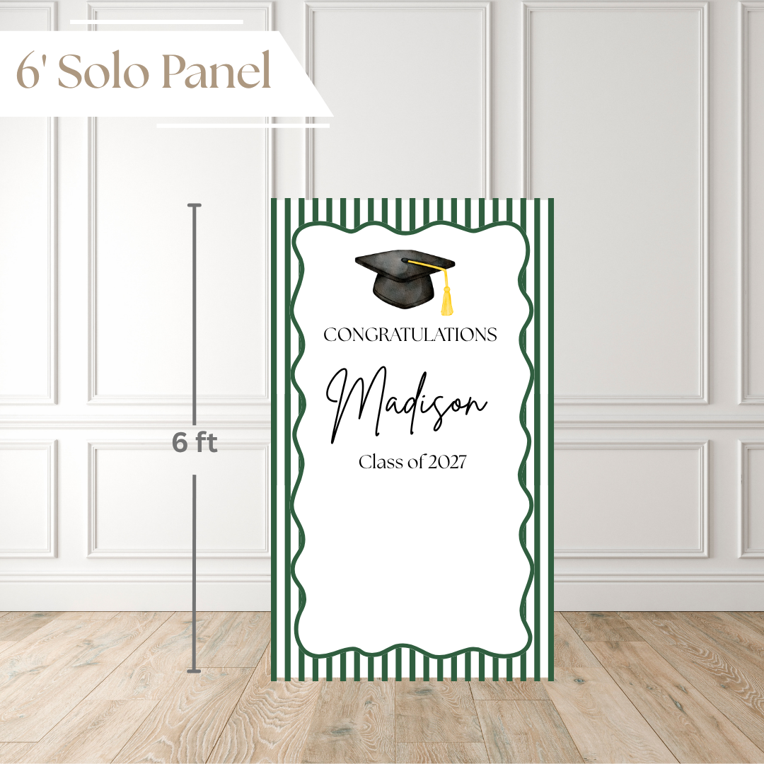 A personalized graduation solo panel sign with green and white stripes, a black graduation cap illustration, and the text 'Congratulations Madison Class of 2027,' shown standing on a wood floor against a white paneled wall with a 6 ft height marker and '6' Solo Panel' label.