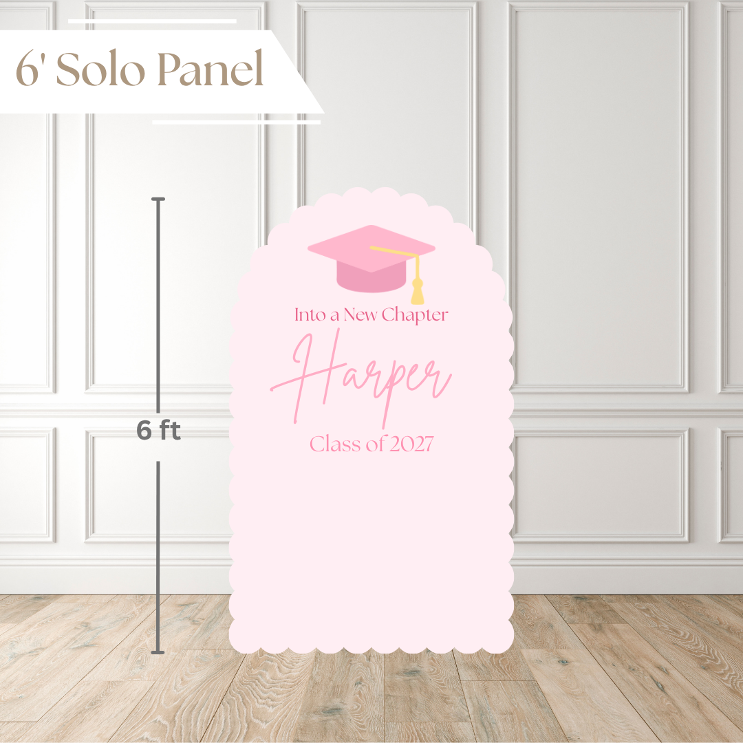 A pink graduation solo panel sign displayed on a light wood floor against a white paneled wall. The sign features a pink graduation cap illustration and customizable text reading 'Into a New Chapter Harper Class of 2027'.