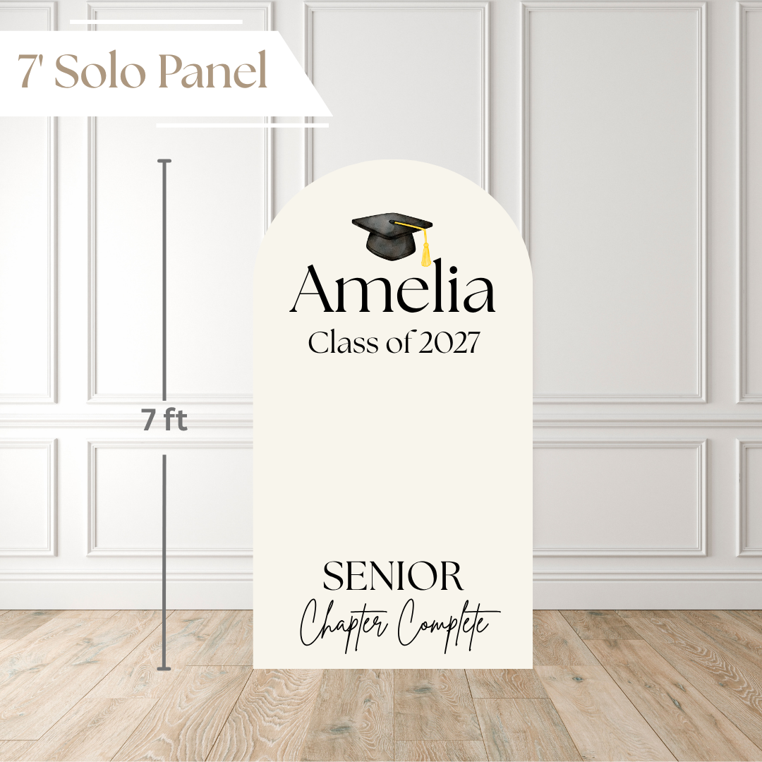 A personalized graduation solo panel sign shown in a room setting with a white paneled wall and wood floor. The sign is cream-colored with a black arch top, featuring a black graduation cap icon, the name 'Amelia', 'Class of 2027', and 'SENIOR Chapter Complete'.