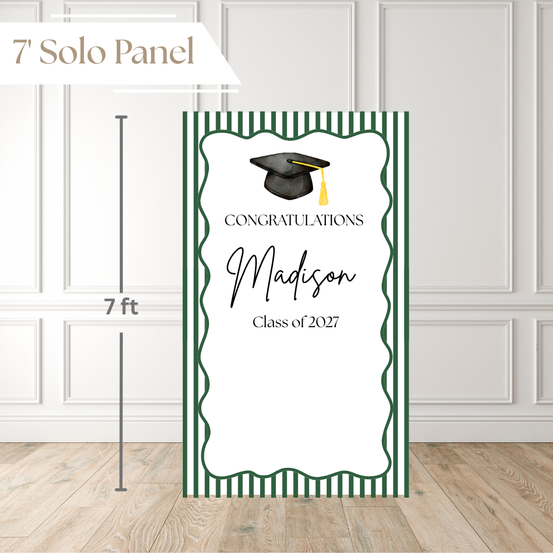 A personalized graduation congratulations sign shown standing on a wood floor against a white paneled wall. The sign has green and white vertical stripes, a black graduation cap illustration, and the text 'CONGRATULATIONS Madison Class of 2027.