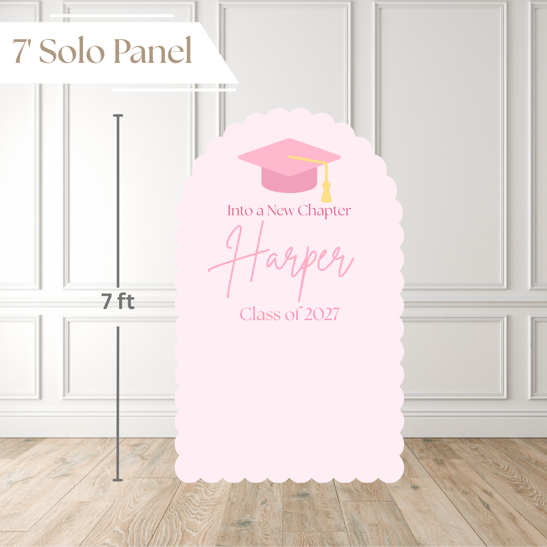 A 7-foot tall light pink graduation solo panel with scalloped edges, featuring a pink graduation cap illustration and customizable text reading 'Into a New Chapter Harper Class of 2027'. The image includes a white paneled wall and wood floor background, with a dimension marker indicating 7 feet.