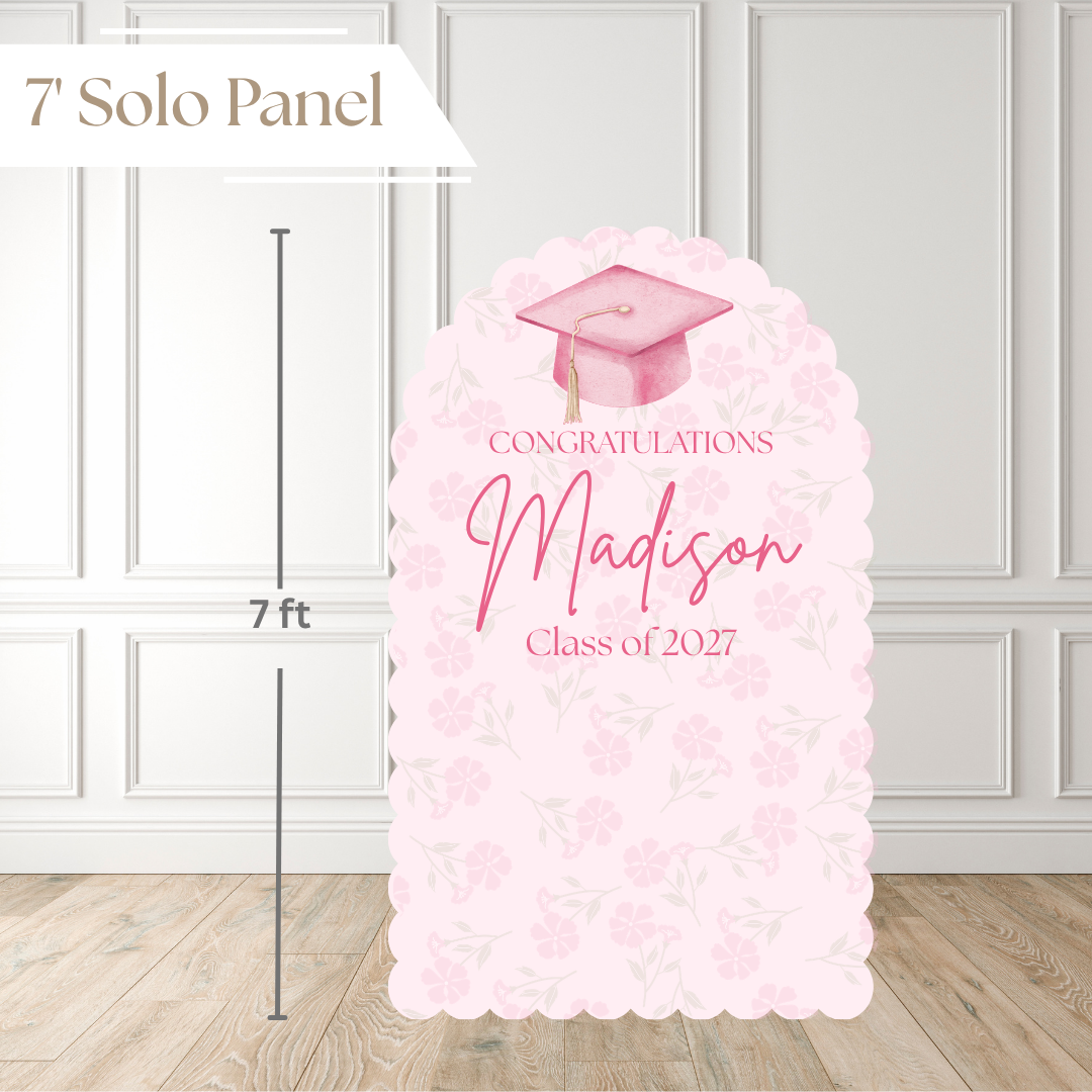 A 7-foot tall graduation solo panel with a scalloped border, featuring a pink watercolor graduation cap illustration and the customizable text 'Congratulations Madison Class of 2027' on a light pink floral pattern. The image includes a white paneled wall and wood floor background with a dimension line indicating the height.